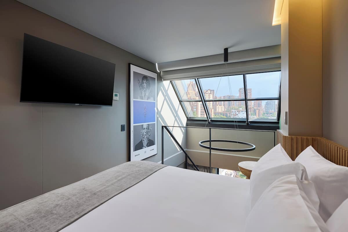 A modern hotel room with a large bed, a flat-screen TV mounted on the wall, a window with a view of the city, and a small table with a circular glass top.