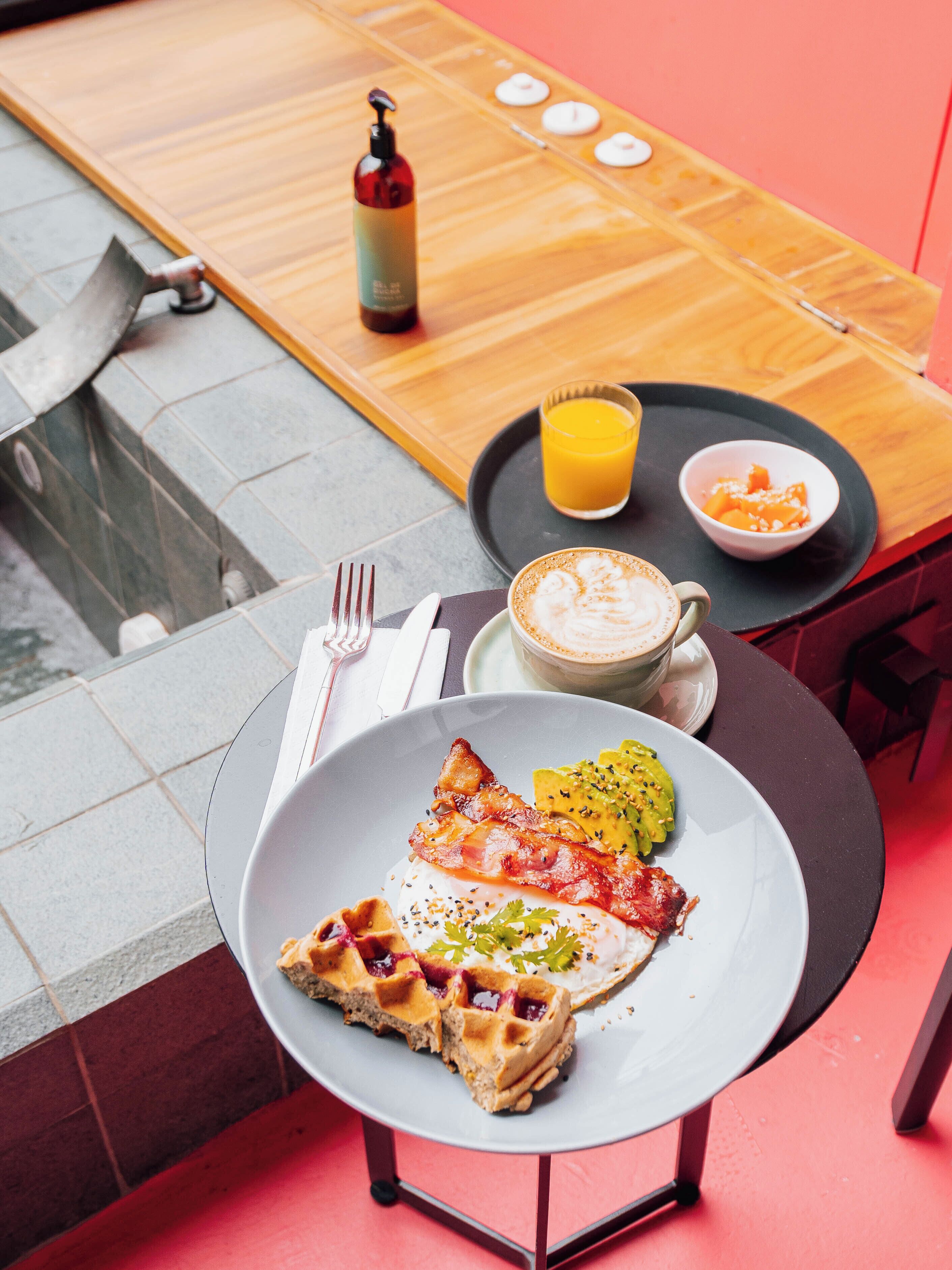 Un desayuno con waffles, tocino y fruta en un plato, una taza de café y un vaso de zumo de naranja sobre una mesa.