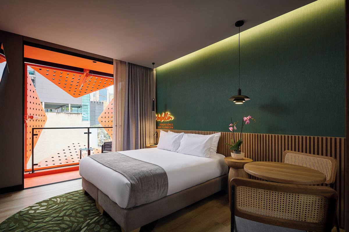 A hotel room with a bed, a small round table, a chair, a potted plant, and a view of a building through a window with a curtain.