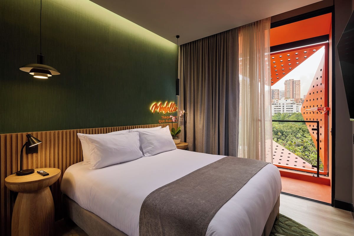 A hotel room with a bed, a nightstand, a neon sign, a window with curtains, and a view of buildings.