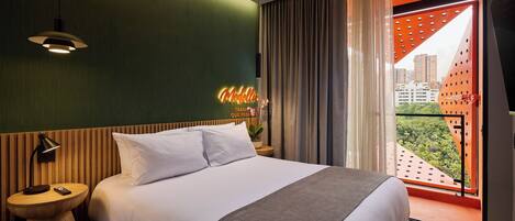 A hotel room with a bed, a nightstand, a neon sign, a window with curtains, and a view of buildings.