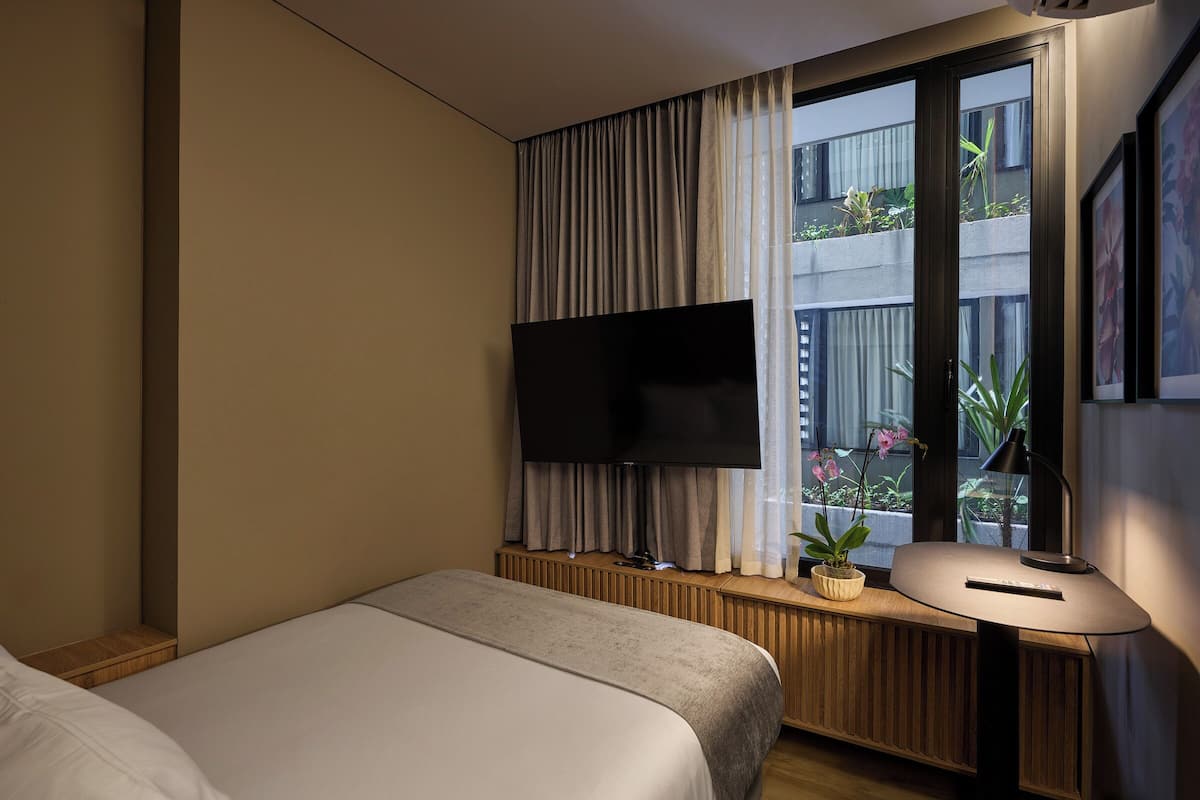 A hotel room with a bed, a TV mounted on a wall, a window with curtains, a bedside table with a lamp, and a small potted plant.