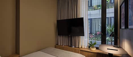 A hotel room with a bed, a TV mounted on a wall, a window with curtains, a bedside table with a lamp, and a small potted plant.