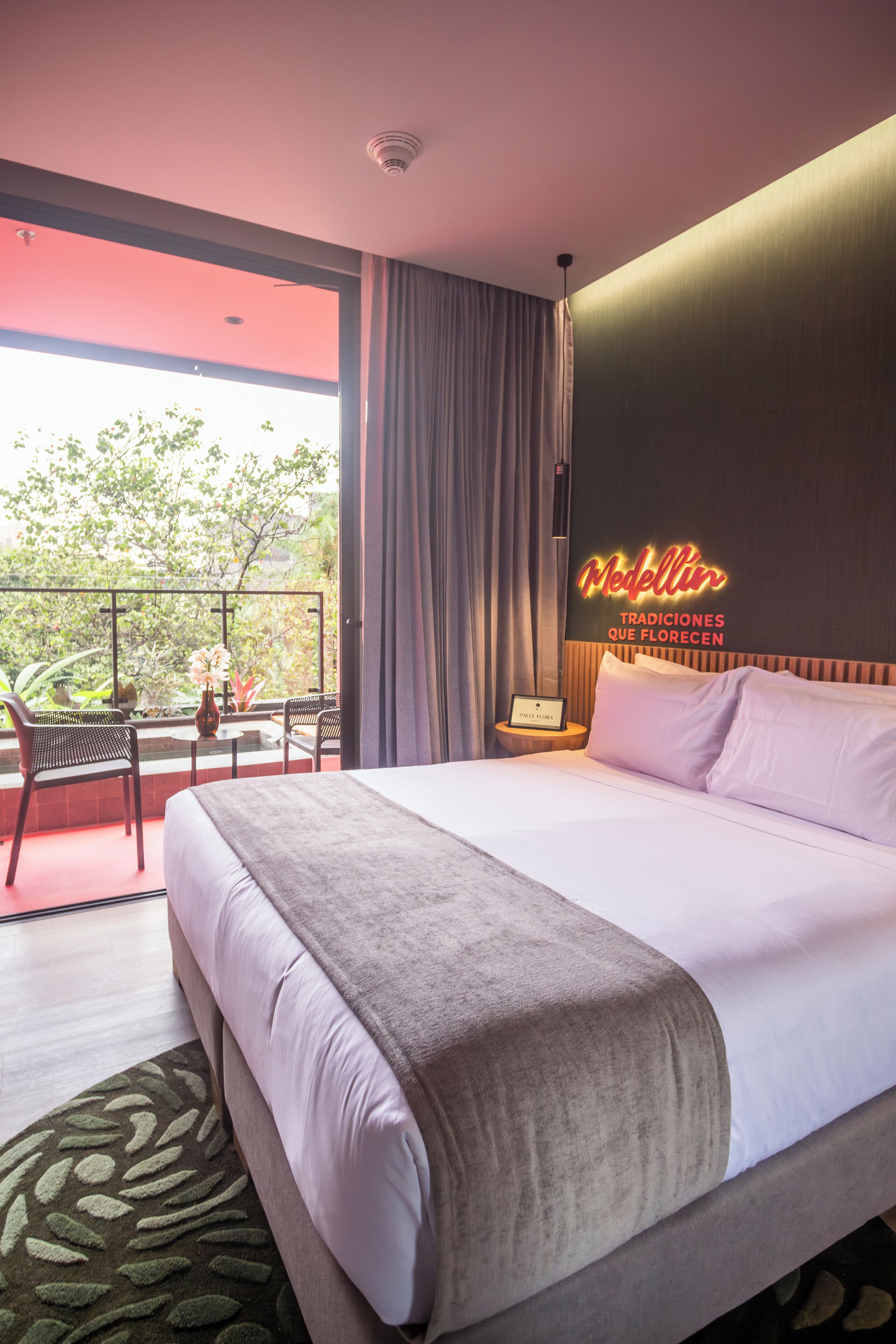 A hotel room with a bed, a balcony with a view, a neon sign, and a small table with a lamp.