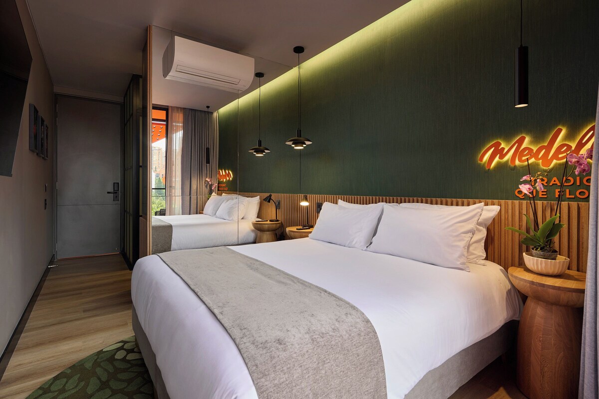 Habitación de hotel con dos camas, un letrero de neón, lámparas colgantes, una planta en maceta y un cabecero de madera.