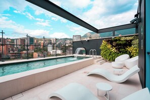 Una piscina en la azotea con una cascada, sillas de descanso blancas y vistas de la ciudad.
