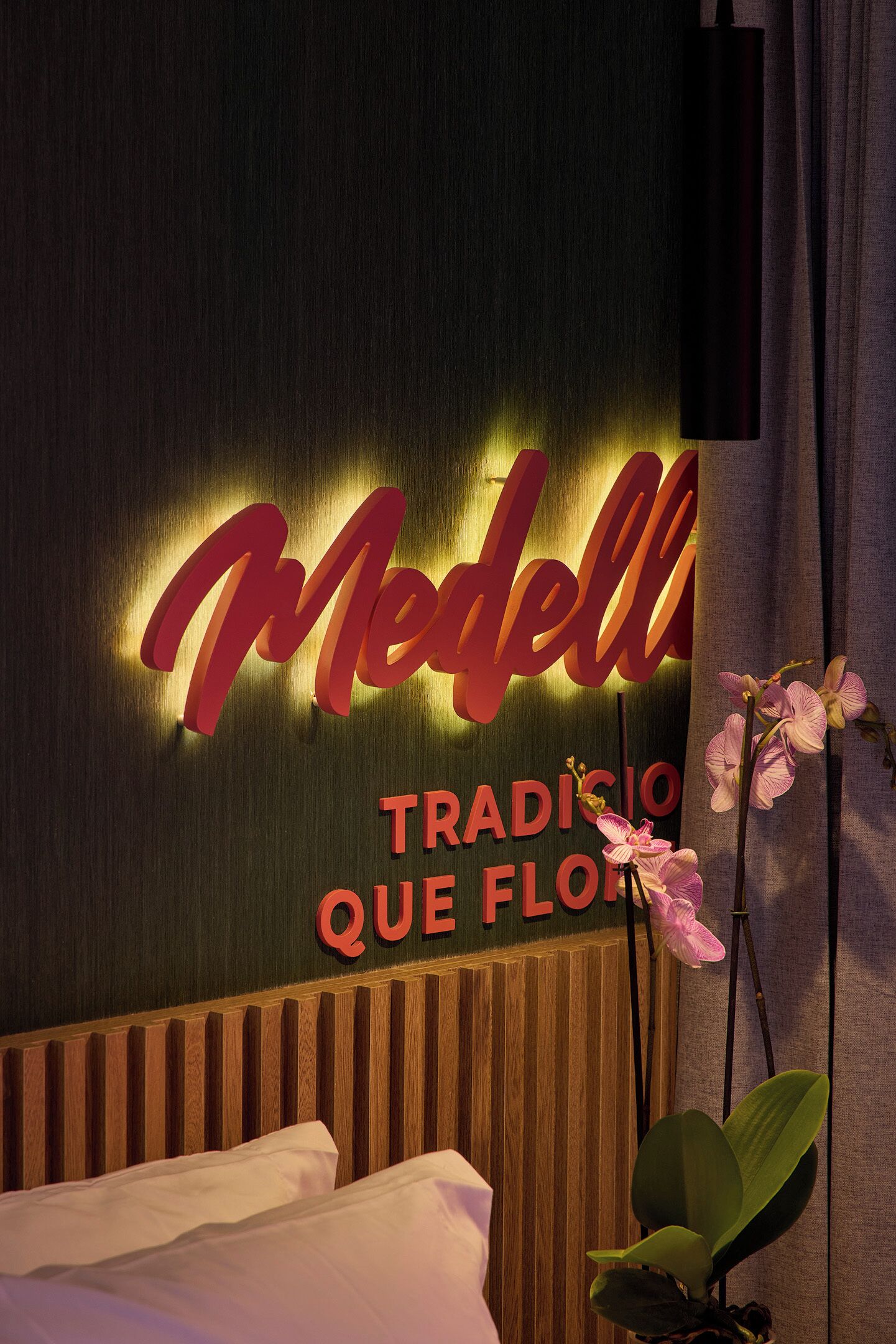 A hotel room with a bed, a wall with a 'Modell' sign, and a potted orchid.