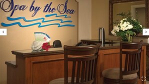 Un mostrador de recepción con un letrero que dice "Spa by the Sea", sillas de madera, un jarrón con flores y un espejo en la pared.