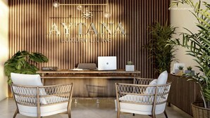 Un mostrador de recepción con un fondo de madera y el nombre 'AYTANA LUXURY LIVING'. Hay dos sillas de mimbre frente al mostrador.