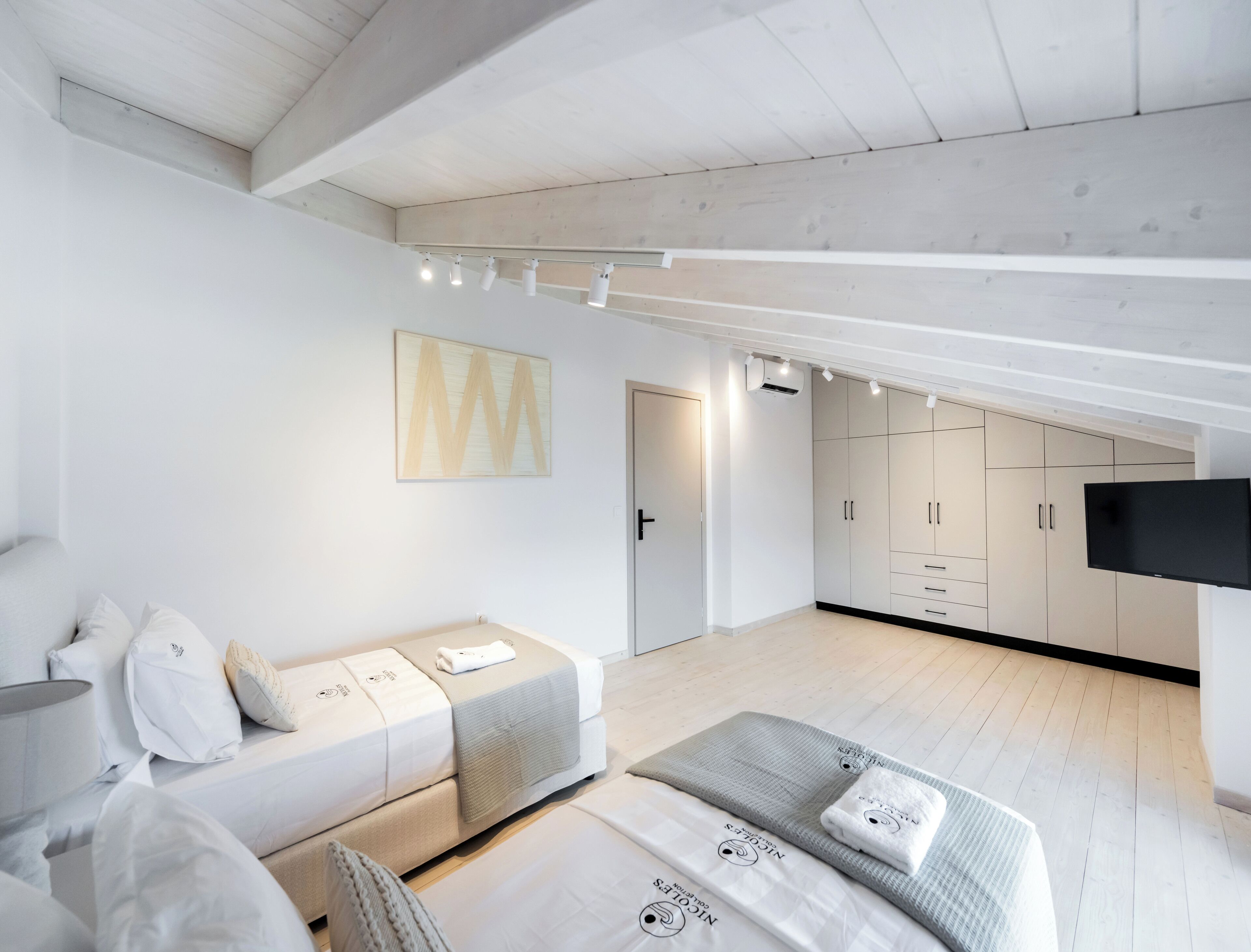 A modern bedroom with a loft-style ceiling, a bed with a grey headboard, a seating area with cushions, a wardrobe, and a flat-screen TV.
