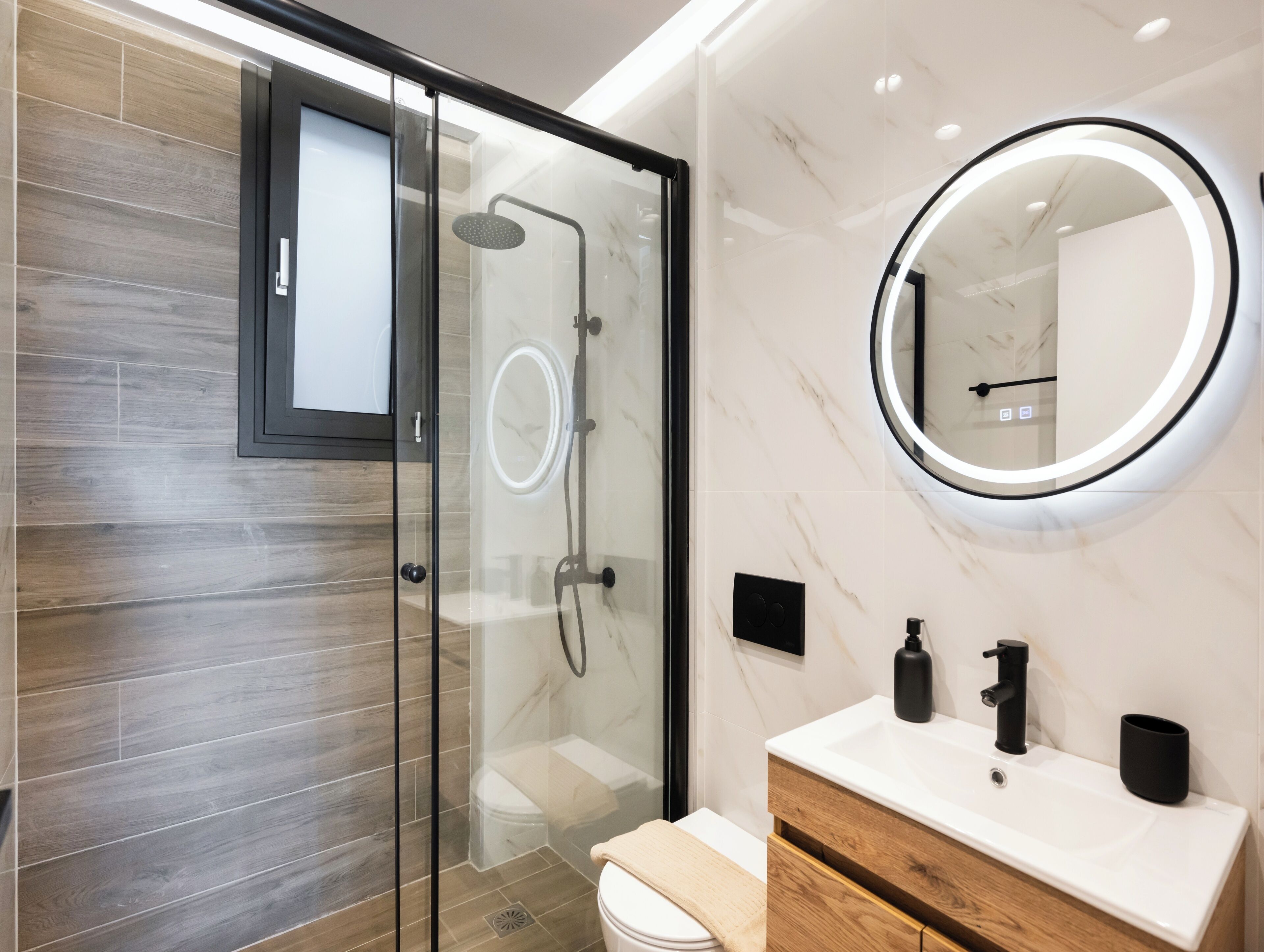 A modern bathroom with a glass-enclosed shower, a vanity with a marble countertop, and a wall-mounted mirror with a circular light.