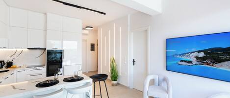 A modern kitchen with white cabinets, a marble backsplash, and a built-in oven. There are two white bar stools, a potted plant, and a large flat-screen TV mounted on the wall.