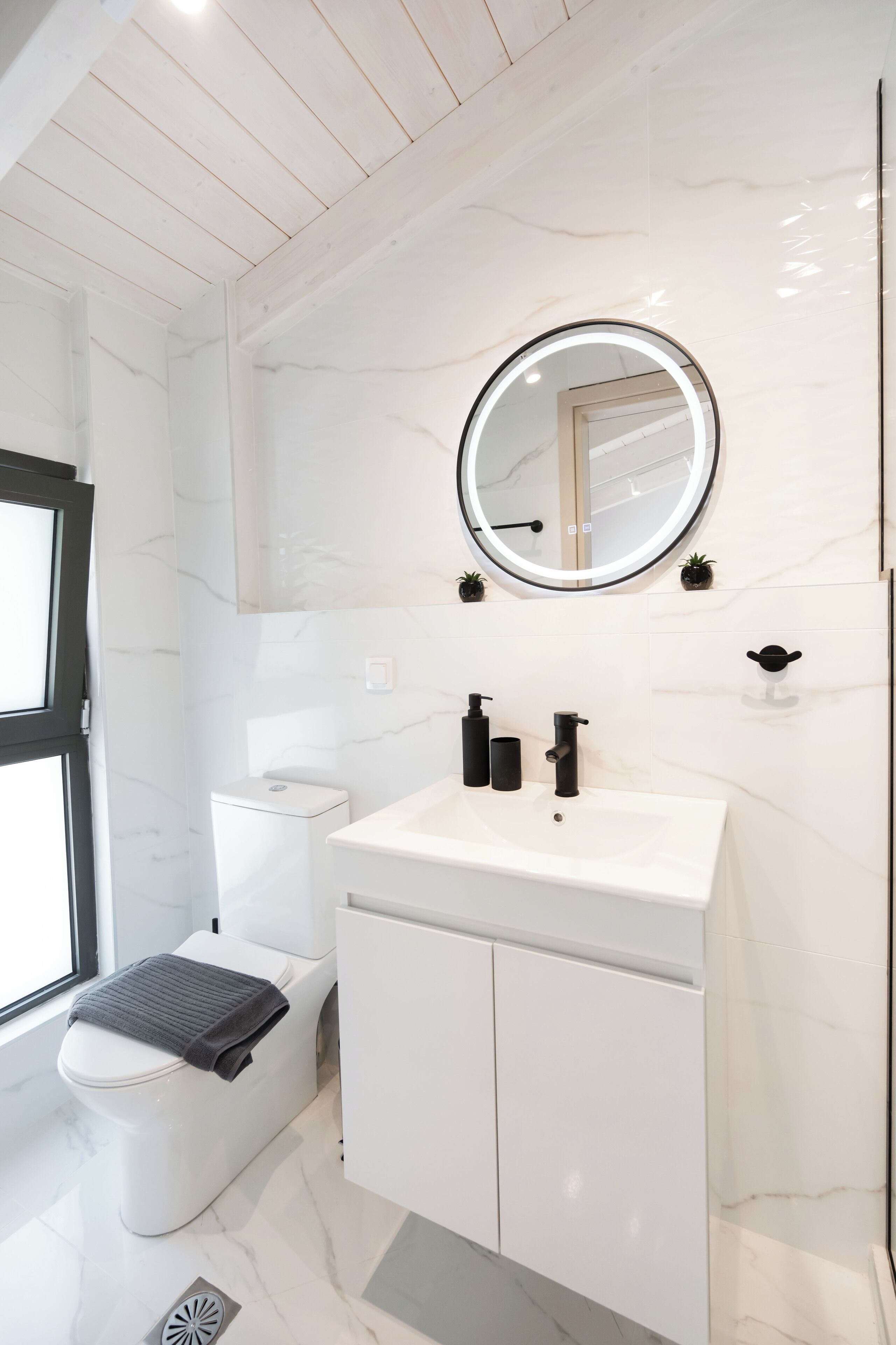A modern bathroom with a white marble floor, a white toilet, a white sink with black faucets, and a round mirror with a light.