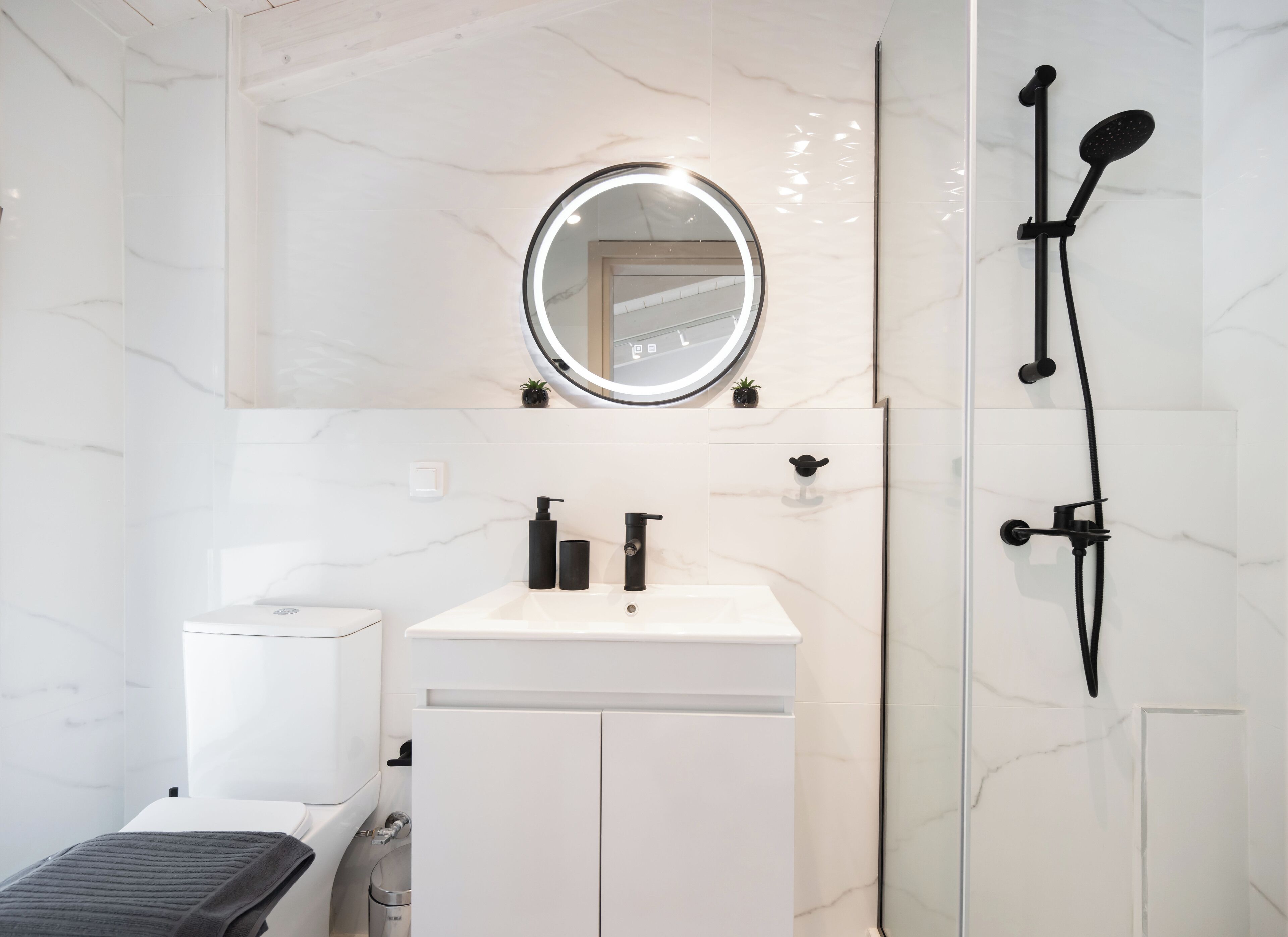 A modern bathroom with a white marble sink, a round mirror with built-in lighting, a handheld showerhead, and a toilet.
