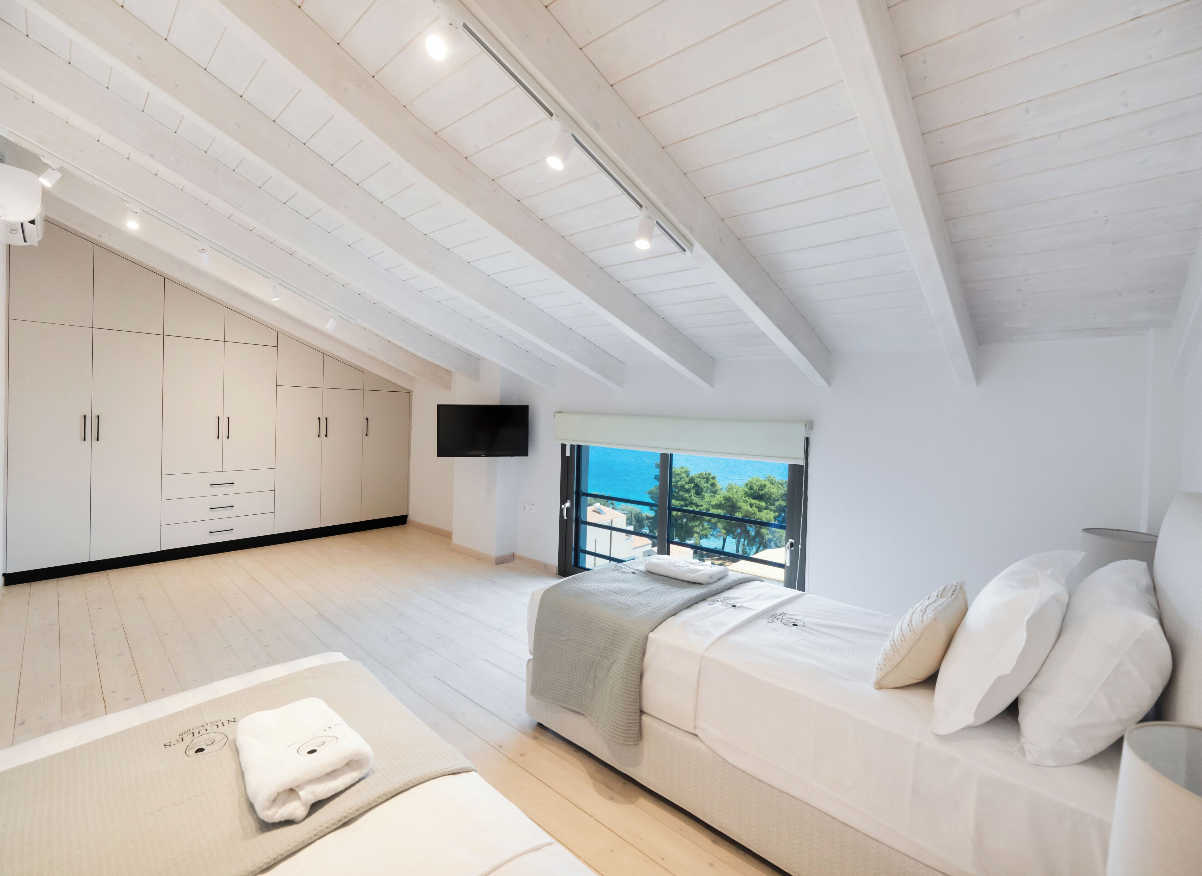 A modern bedroom with a large bed, a TV, and a balcony with a view of the sea.