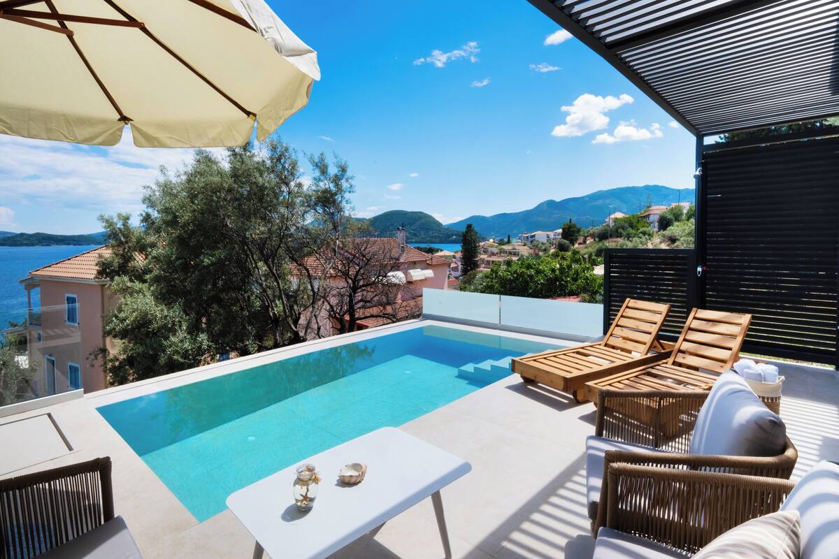 Un área de piscina en la azotea con vista despejada al mar y a las montañas, sillones de madera para tomar el sol y una mesa con un florero y una concha.