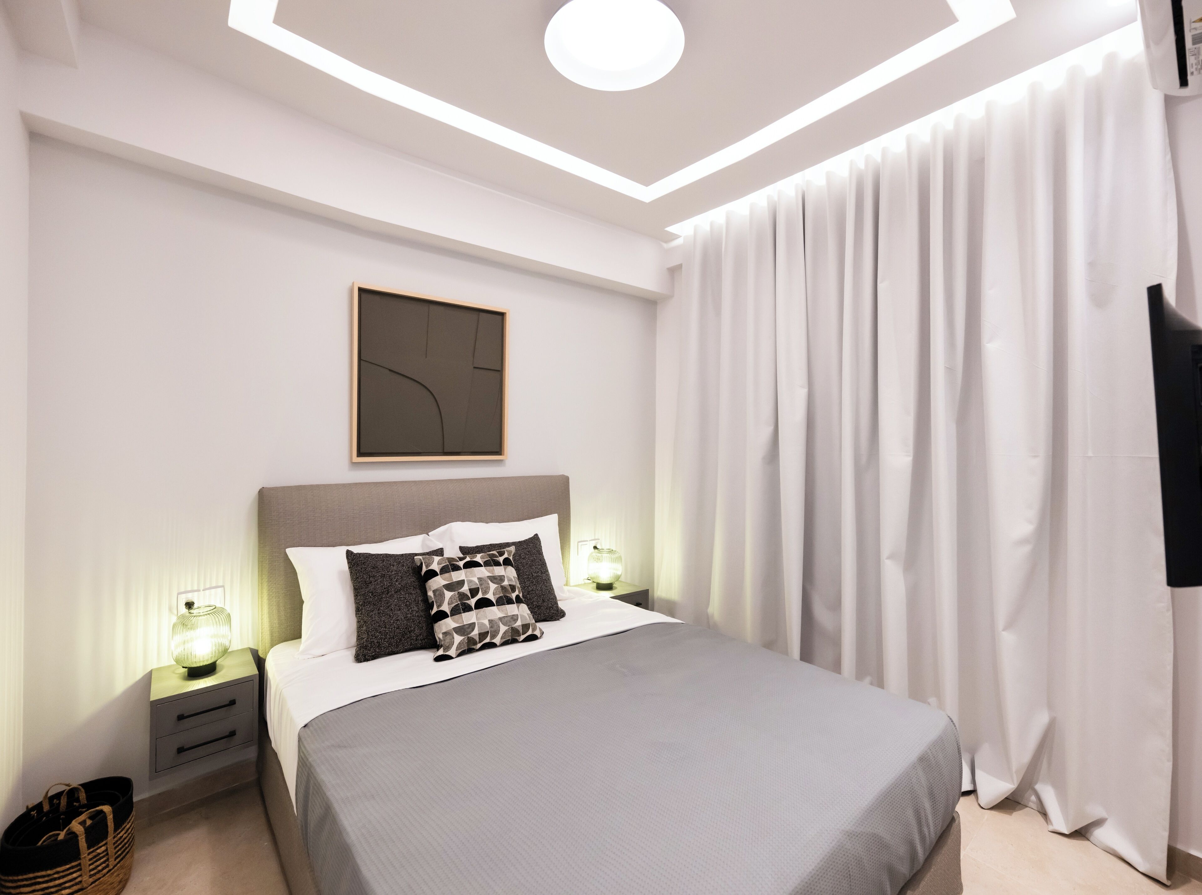 A modern bedroom with a large bed, bedside tables with lamps, a framed abstract artwork, and a large window with curtains.