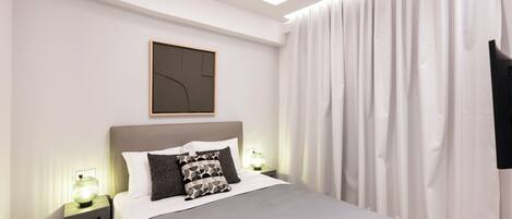 A modern bedroom with a large bed, bedside tables with lamps, a framed abstract artwork, and a large window with curtains.