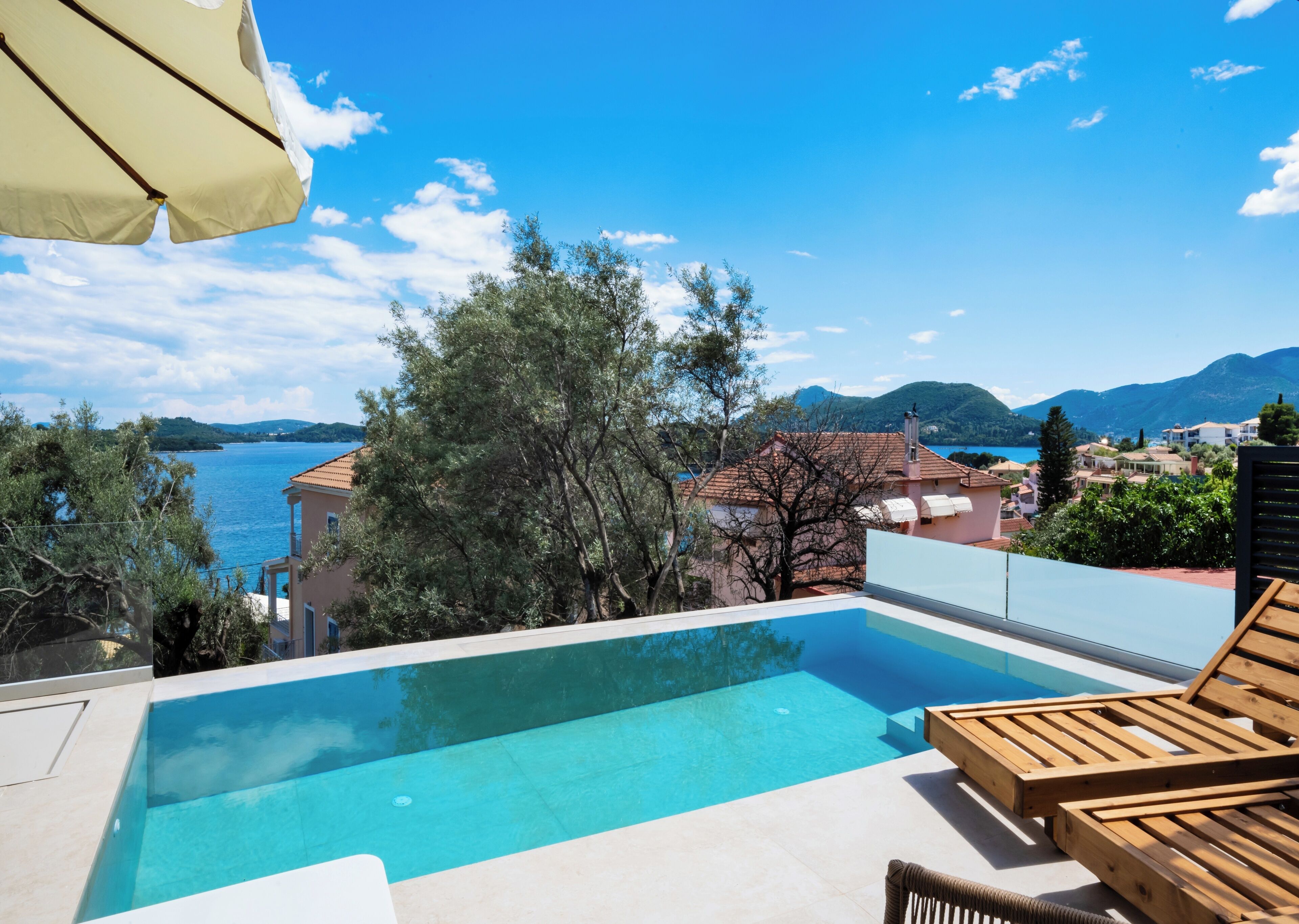 Piscina infinita com vista para um lago e montanhas, cadeiras de madeira para relaxar e um guarda-sol bege.
