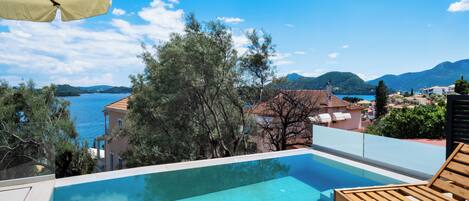 An infinity pool with a view of a lake and mountains, wooden lounge chairs, and a beige umbrella.