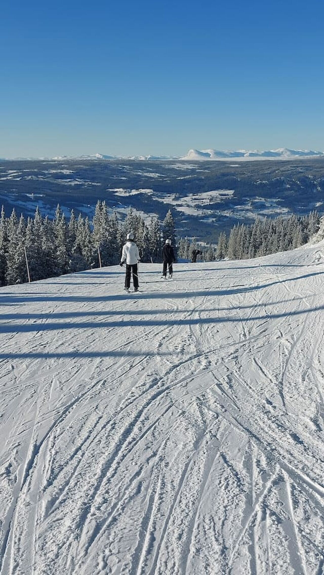 Pista de esquí nevada con esquiadores y una vista despejada de montañas distantes.
