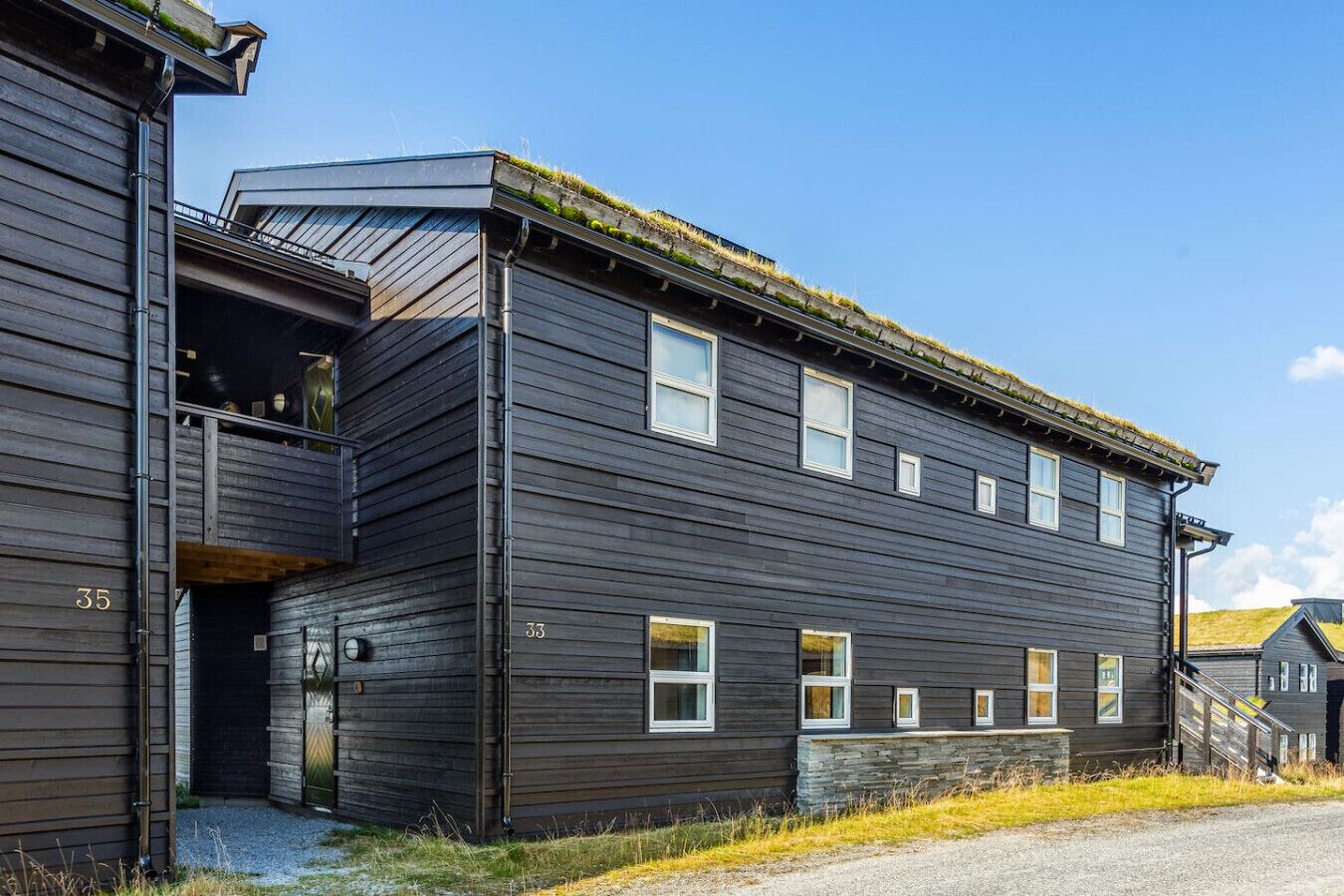 Un edificio de dos pisos con fachada de madera oscura, con los números 33 y 35, que presenta un balcón y un techo plano cubierto de césped.