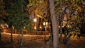 Una terraza de madera con un banco, rodeada de árboles e iluminada por luces cálidas.