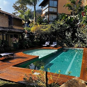A swimming pool with a wooden deck, surrounded by lush greenery and a building with a tiled roof.