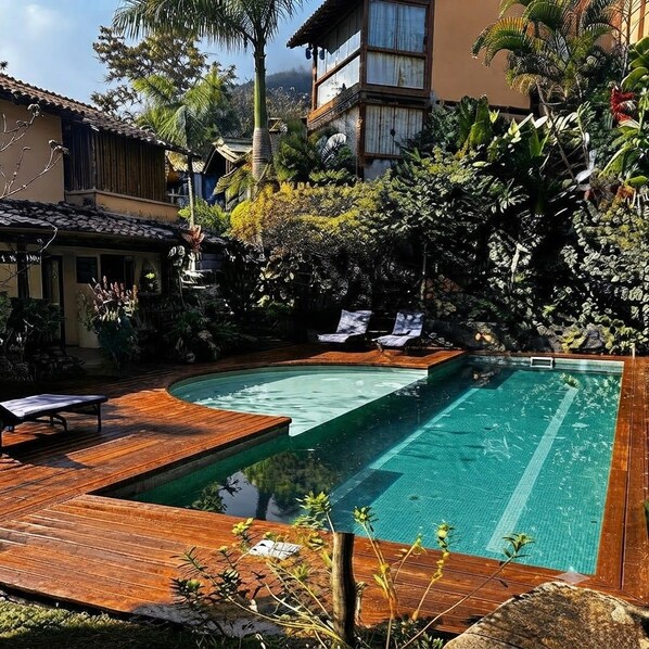 A swimming pool with a wooden deck, surrounded by lush greenery and a building with a tiled roof.