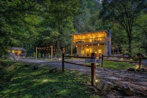 A two-story wooden cabin with a balcony, surrounded by trees and a gravel path.