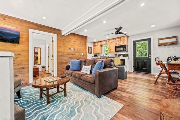 A cozy living room with a wooden floor, a brown leather sofa, a wooden coffee table, and a blue patterned rug.