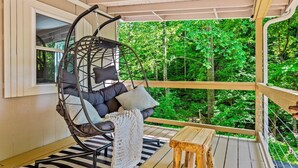 A wicker chair with cushions and a blanket, a wooden table, and a view of a forested area through a balcony railing. - NEW Listing 🔥 Hot Tub • Mountain View • Fire Pit • EV Charger (Sevierville)