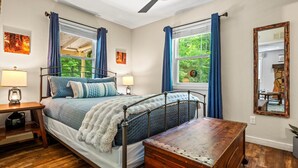 Une chambre à coucher avec un lit, des lampes de chevet, une commode en bois, un ventilateur de plafond et des fenêtres avec des rideaux.