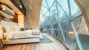 Moderni makuuhuone, jossa on suuri sänky, pyöreä seinäkello ja näkymä ulos geometrisesti muotoillun ikkunan kautta.