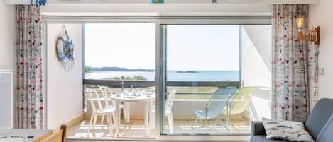 A room with a sliding glass door leading to a balcony with a view of the sea. There is a table with chairs and a sofa.