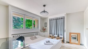 Une chambre à coucher comprenant un lit, un bureau, une chaise, une fenêtre donnant sur un espace verdoyant, un luminaire au plafond et un miroir.