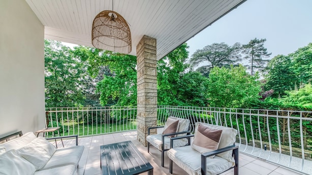 Un balcon doté d’un canapé blanc avec des coussins, d’une petite table et de deux fauteuils également pourvus de coussins. Le balcon est agrémenté d’une balustrade blanche et offre une vue sur une végétation luxuriante.