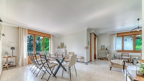 Une pièce spacieuse comprenant une table à manger et des chaises, un canapé et un fauteuil avec une petite table. On y trouve des fenêtres avec des rideaux, un radiateur et une lampe.