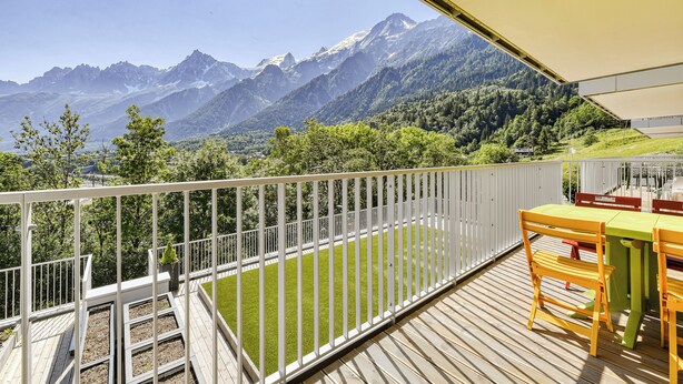 A deck with a view of snow-capped mountains, a white railing, and a table with chairs.