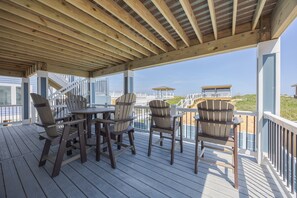 A covered outdoor deck with a table and chairs, overlooking a beach and buildings.