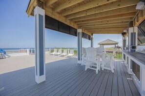 A covered outdoor deck with white chairs and tables, overlooking a beach. - 5389- Heavenly Sunrise - Oceanfront Estate for 32 | Pool & Theater (Kill Devil Hills)