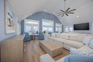 A spacious living room with a high ceiling, large windows, a ceiling fan, and a flat-screen TV mounted on the wall. - 5389- Heavenly Sunrise - Oceanfront Estate for 32 | Pool & Theater (Kill Devil Hills)