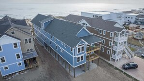 A coastal building with a large bay window, a covered porch, and a staircase leading to a balcony. - 5389- Heavenly Sunrise - Oceanfront Estate for 32 | Pool & Theater (Kill Devil Hills)