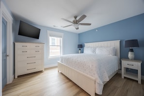 Un dormitorio con cama, cómoda, televisor, ventilador de techo y ventana.