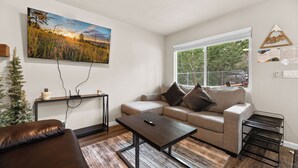 A living room with a sofa, a coffee table, a TV mounted on the wall, and a window with a view of trees. - The Maple Lane Hideaway (Sugarloaf)