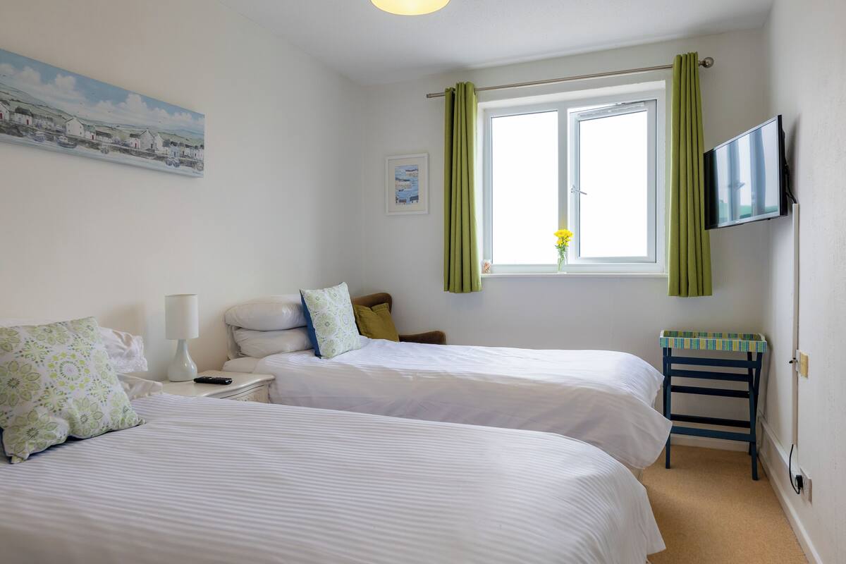 A hotel room with two beds, a TV, a window with curtains, a painting on the wall, and a small table with a lamp.