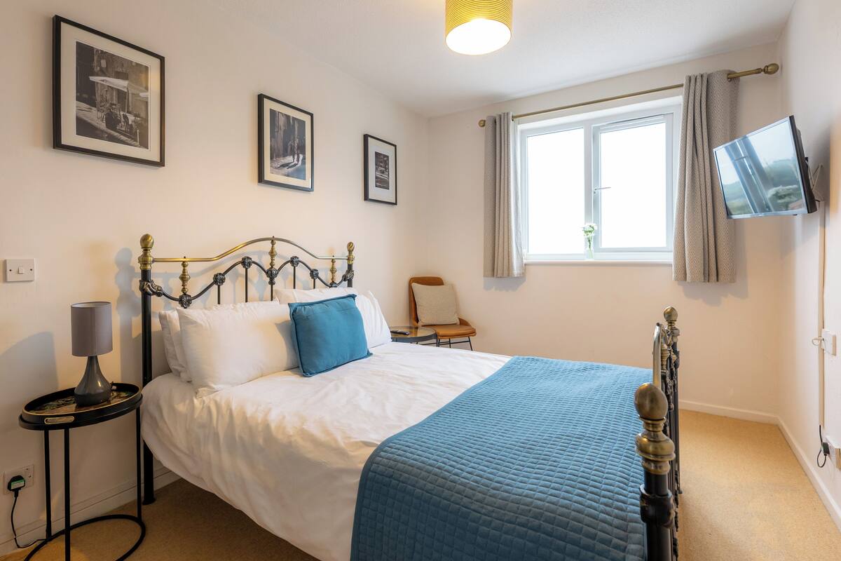 A neatly made bed with a blue quilt, a bedside table with a lamp, and a television mounted on the wall.