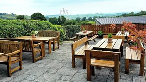 An outdoor seating area with wooden tables and benches, surrounded by greenery and a view of the countryside. - Hawk and Buckle Inn (Llannefydd)