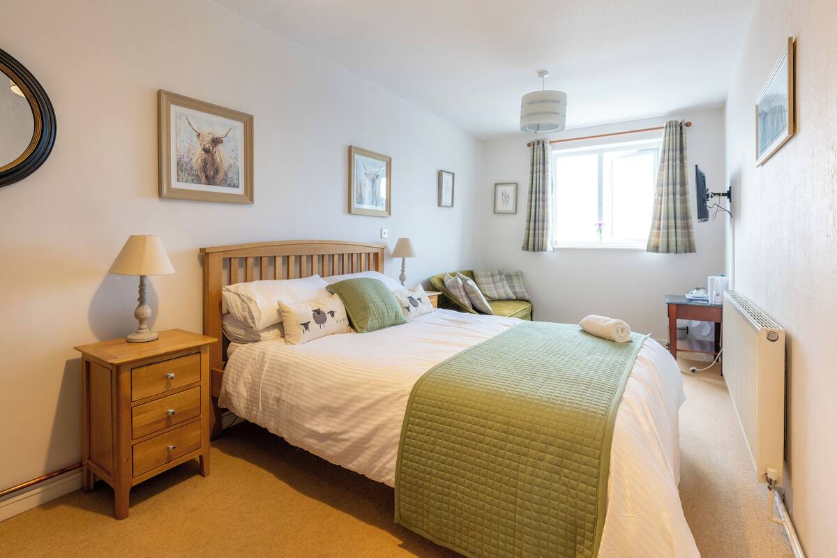 A bedroom with a bed, a nightstand, a lamp, a window with curtains, and wall-mounted pictures.