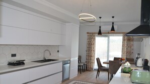 A modern kitchen with white cabinets, a dishwasher, a dining area with a table and chairs, and a green toaster. - VILLA WITH LAKE AND FOREST VIEW IN DALYAN2 (Akcakavak)
